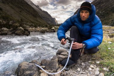 male backpacker in a blue down jacket filters drinking water from a mountain stream in the Cordillera Blanca in the Andes in Peru clipart