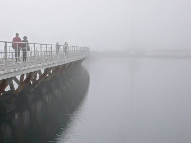 pedestrians crossing a long wooden bridge over calm water on a foggy and misty morning