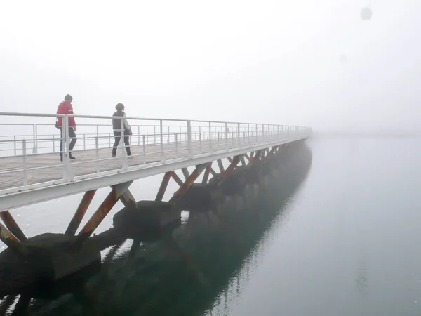 Pedoni che attraversano un lungo ponte di legno su acque calme in una mattina nebbiosa e nebbiosa — Foto Stock
