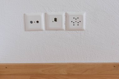 Beyaz alçı duvar ve ahşap zemin üzerinde televizyon, internet, telefon ve elektrik prizlerinin yakın görüntüsü