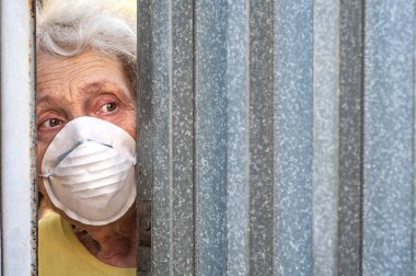 Maskeli ürkek bir büyükanne evinin kapısından çitteki boşluktan dışarı bakıyor. İzolasyon. Coronavirus covid-19. Yaşlı insanları karantinaya alın. Evde kal. Agorafobi, zihinsel bozukluk.