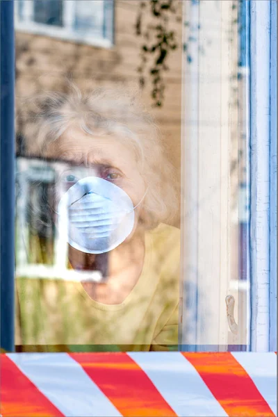 A sad elderly wrinkled woman is looking through the window in the respiratory mask of her house. Isolation of the elderly coronavirus covid-19. Quarantine and isolation to keep old people. Guard tape
