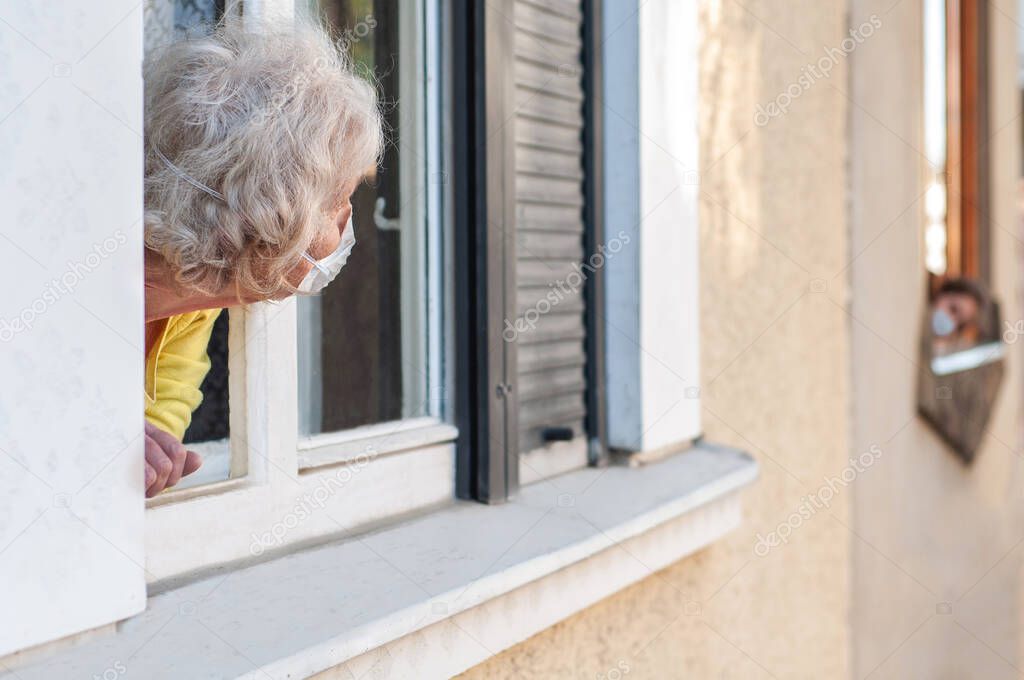 Grandmother and neighbor child in protective masks communicate through the windows of their apartments. Keep a distance. Coronavirus covid-19. Quarantine and isolation. Stay at home.