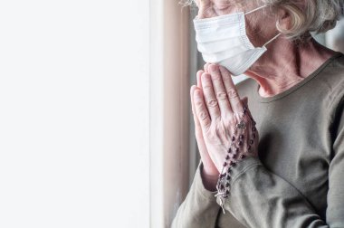 Pencerenin yanında tıbbi solunum maskesi takmış yaşlı bir kadın tespih ve haçla dua ediyor. Din. İnsan sağlığı için dua etmek. Coronavirus covid-19 salgını. Karantina ve izolasyon.
