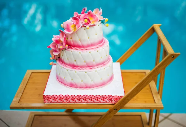 A large pink and white holiday cake decorated with flowers on a wooden tray on a background of water. Side view.