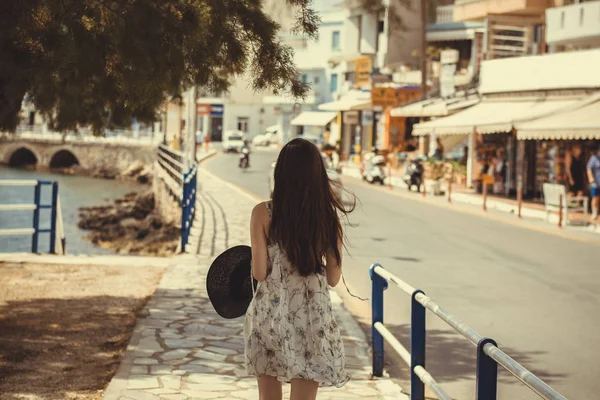 Uma jovem com chapéu preto na mão caminha ao longo do cais da cidade de Rethymno, Grécia. Menina em um chapéu de palha andando ao longo do passeio. Vista de trás — Fotografia de Stock