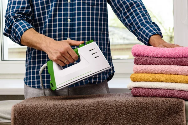 Hombre plancha ropa y toallas sobre tabla de planchar con plancha humeante blanco-verde . — Foto de Stock