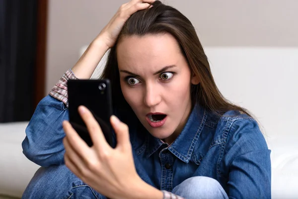 Young woman holding his head and looks to the smartphone. Sitting on the floor in shock. — Stock Photo, Image