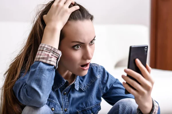 Young woman holding his head and looks to the smartphone. Distressed by what he saw — Stock Photo, Image