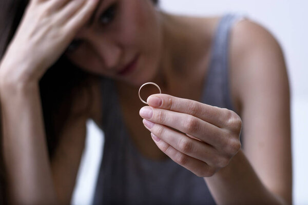 Disappointed wife depressed after divorce, holding a wedding ring sitting on a white sofa