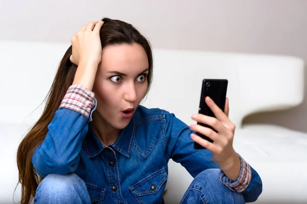 Mujer joven sosteniendo su cabeza y mira al teléfono inteligente. Sentado en el suelo en shock . —  Fotos de Stock