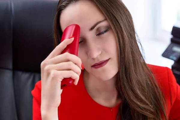 Mulher de negócios atraente de cabelos escuros em terno vermelho e telefone vermelho na mão sofrendo de dores de cabeça. O conceito de trabalho duro no escritório. monocromático . — Fotografia de Stock