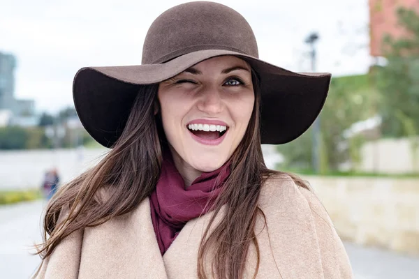 Happy woman in a brown hat and beige coat, winking one eye at the camera and smiles widely with white teeth. Pick me.