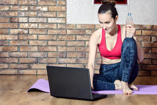 Attractive middle-aged brunette takes a break from exercises while sitting on a gym Mat, drinking water, and overcame instructional videos online. — Stock Photo, Image