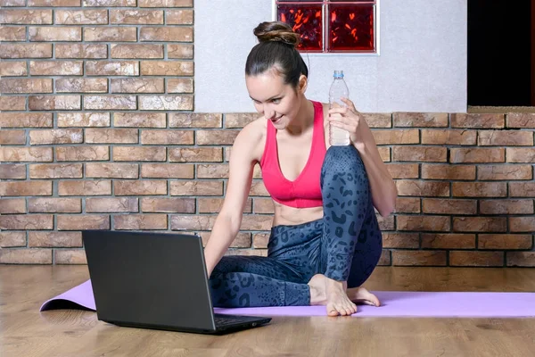 Αστείο κορίτσι σε αθλητικά ενδύματα παίρνει ένα διάλειμμα από τις ασκήσεις ενώ κάθεται στο πάτωμα και βλέποντας το κοινωνικό δίκτυο στο laptop με ένα μπουκάλι νερό στο χέρι — Φωτογραφία Αρχείου