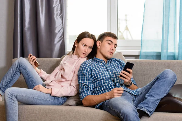 Νεαρό ζευγάρι κάθεται στον καναπέ και τη χρήση των smartphones, η σύζυγος υποψιάζεται το σύζυγό κατάσκοποι στο τηλέφωνό του — Φωτογραφία Αρχείου