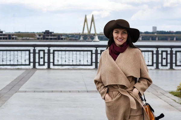茶色の帽子、ベージュのコートの女性が広く白い歯と笑顔のウォーター フロントに沿って歩く幸せ — ストック写真