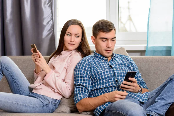 Νεαρό ζευγάρι κάθεται στον καναπέ και χρησιμοποιώντας το smartphone, το κορίτσι spies μέσω ένας τύπος κινητό τηλέφωνο — Φωτογραφία Αρχείου