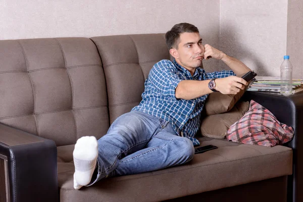 Un joven en jeans, con un control remoto para el aburrimiento de la televisión en la cara cambia el canal — Foto de Stock