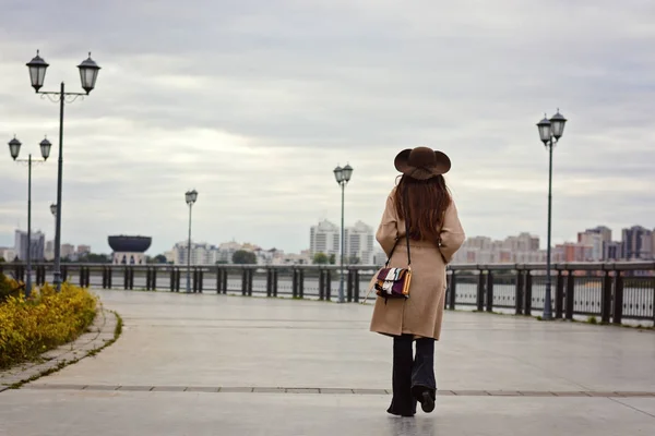 Привлекательная стильная девушка в длинном пальто с поясом и большой шляпой, идущая по набережной. Осенние прогулки на свежем воздухе — стоковое фото