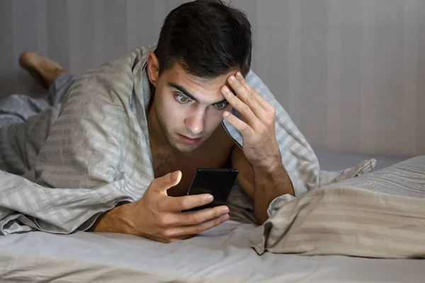 Giovane uomo frustrato e stressato è in ritardo. Si sta svegliando, guarda lo smartphone ed è scioccato. Il concetto di un ritmo di vita moderno, l'insonnia — Foto Stock