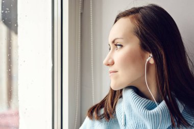 Yağmur, bulutlu ve soğuk hava odasında pencere bir smartphone ile kulaklıklar müzik dinlemek mavi yün kazaklı genç esmer kadın.