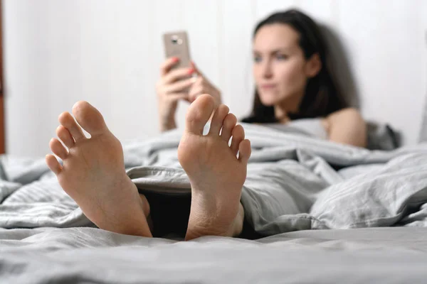 Una giovane donna si trova a letto e comunica nei social network tramite smartphone. Concentrati sui piedi. profondità di campo poco profonda — Foto Stock