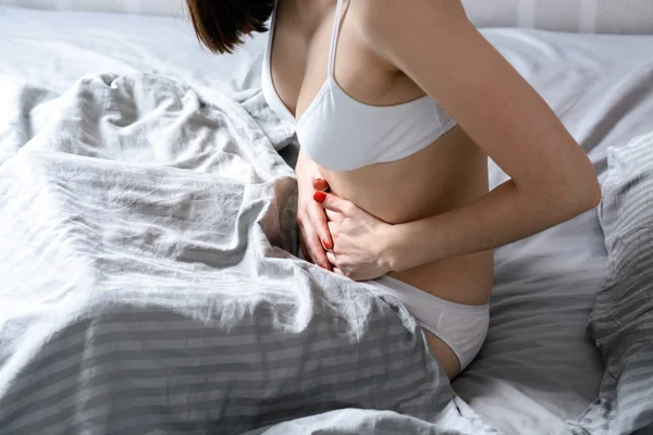 Молодая женщина, держащая живот, сидящий в постели, боль в животе — стоковое фото
