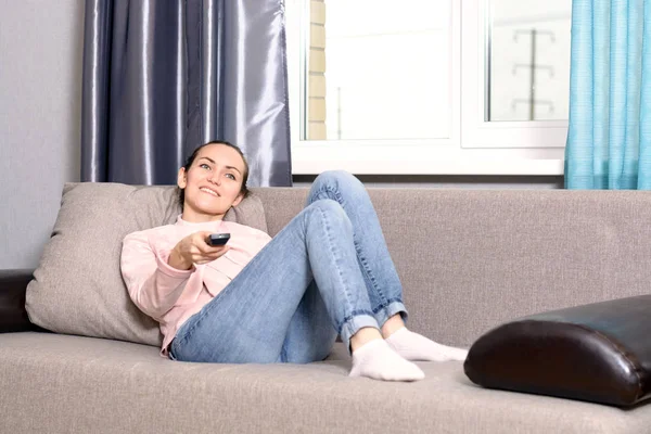 Young woman lying on the sofa with the remote control of the TV and watching a movie — Stock Photo, Image