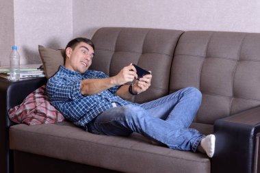 Yazdırma bir kafeste gömlek giymiş bir kıl adamın görüntü Excited, kot pantolon ve beyaz çorap, Divan ev ve smatphone oynarken yatıyor. Oyun hakkında tutkulu