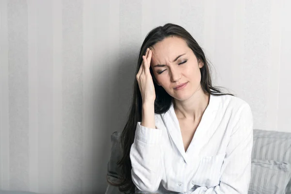 Retrato de una joven morena en pijama sentada en la cama, dolor de cabeza, migraña —  Fotos de Stock