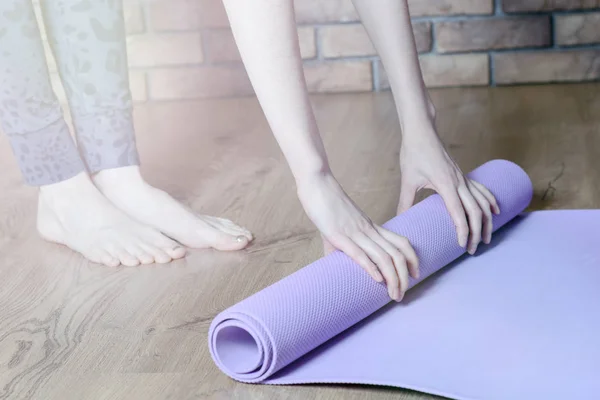 A barefoot woman twists a purple yoga Mat and fitness on the parquet floor — Stock Photo, Image