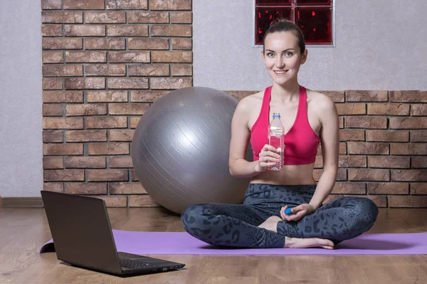 Μια νεαρή γυναίκα σπορ blogger είναι ανάπαυση μετά από μια προπόνηση σε απευθείας σύνδεση, πόσιμο νερό από ένα πλαστικό μπουκάλι στο yoga Mat. Γυμναστικής στο σπίτι — Φωτογραφία Αρχείου