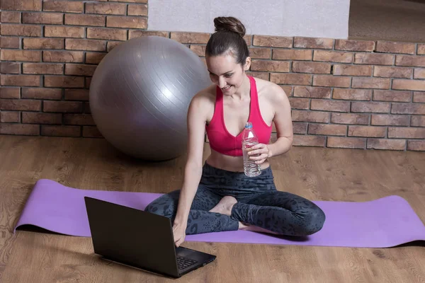 Молода жінка спортивний блогер відпочиває після тренування в Інтернеті, п'є воду з пластикової пляшки на йога мат. Домашня фітнес — стокове фото