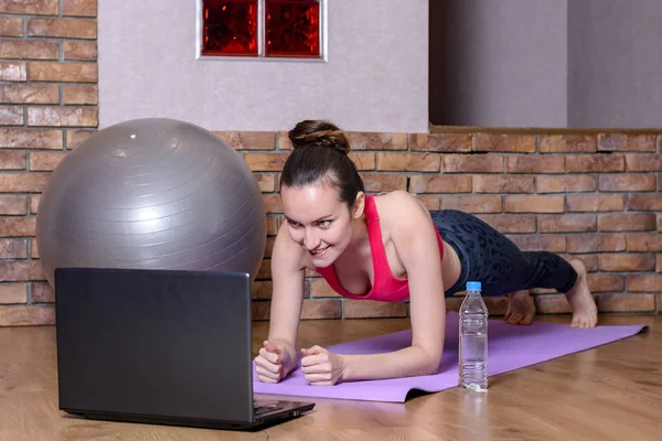 A young woman performs a plank exercise on a purple mat on parquet floor and watching instructional videos on laptop — Stock Photo, Image