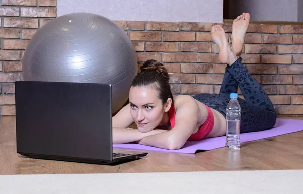 Young girl lying with hands under my head on the yoga Mat and looks at the laptop screen. Fitness at home — Stock Photo, Image