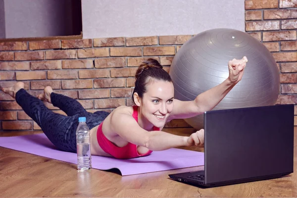 Young smiling woman lying on fitness Mat depicts the flight of a superhero during the warm-up before training online with videos on the laptop. funny home fitness — Stock Photo, Image