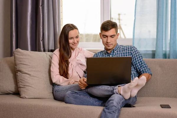 A young couple sitting on the couch in the room and watch the video on the laptop on his knees — Stock Photo, Image