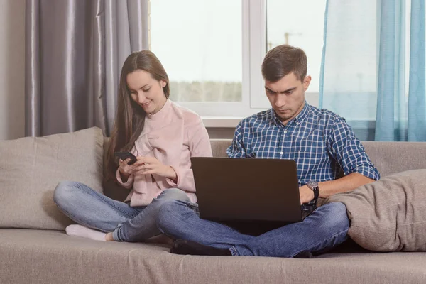 Pareja joven sentada en el sofá de la habitación, ocupada con sus aparatos, problemas de comunicación de la sociedad moderna — Foto de Stock