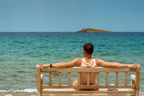 Jovem com braços estendidos, sentado em um banco de madeira na margem do mar Mediterrâneo na ilha de Creta e olhando para o horizonte — Fotografia de Stock