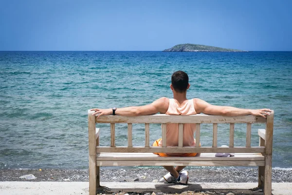 Νεαρό άνδρα με τα χέρια απλωμένα, κάθεται σε ένα ξύλινο παγκάκι στην όχθη της Μεσογείου στο νησί της Κρήτης και κοιτώντας τον ορίζοντα — Φωτογραφία Αρχείου
