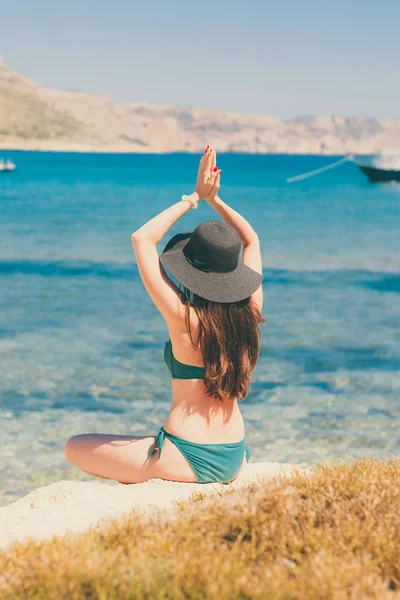 Jovem de fato de banho verde e chapéu preto pratica ioga na praia do mar Mediterrâneo. qigong e liberdade — Fotografia de Stock
