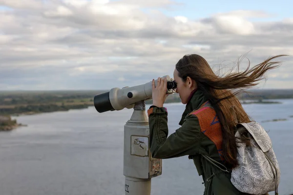 Una giovane viaggiatrice sul ponte di osservazione guardando attraverso il binocolo il panorama della città di Nizhny Novgorod — Foto Stock