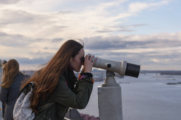 A young woman traveler on the observation deck looking through binoculars at the panorama of the city of Nizhny Novgorod.