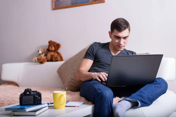 若い男はノート パソコン テーブル ノート パソコンとカメラの横にあるとソファの上に座っています。フリーランスのブログ、今日の若者の概念 — ストック写真