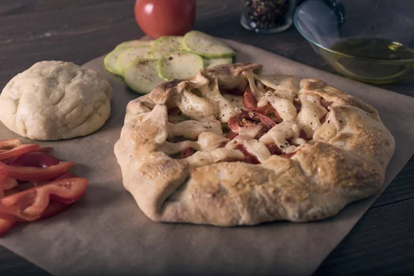 Hausgemüse-Galeta, Pizza auf einem Holztisch im Konturlicht. Zutaten Teig, Tomaten, Zucchini, Mozzarella, Pfeffer, Olivenöl. Backen auf Pergamentpapier — Stockfoto