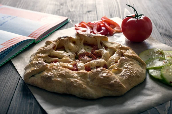 Hausgemüse-Galeta, Pizza auf einem Holztisch im Konturlicht. Zutaten Teig, Tomaten, Zucchini, Mozzarella, Pfeffer, Olivenöl. Backen auf Pergamentpapier — Stockfoto