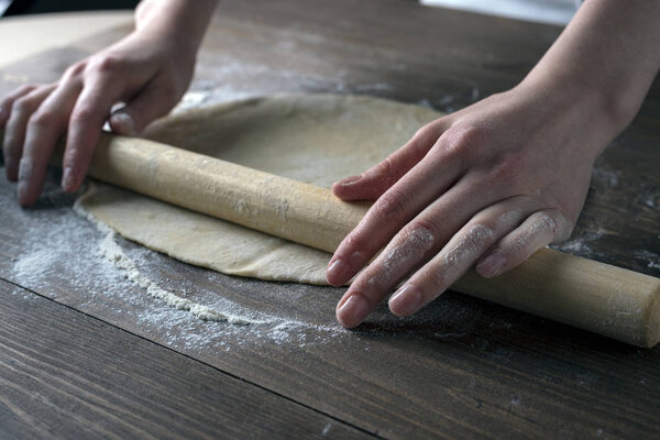 Hands with a rolling pin roll out the dough on a brown wooden table, side light, front view