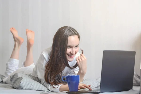 A young woman in pajamas is lying in bed and chatting on social networks through a laptop. Modern youth — Stock Photo, Image