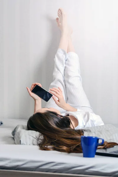 Giovane donna in pigiama sdraiata sul letto, piedi sul muro. Comunicazione nei social network, utilizzare gli smartphone . — Foto Stock